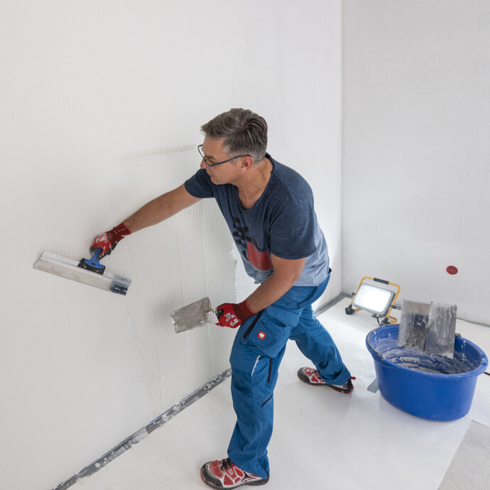Heimwerker zieht im Raum einen weissen, feinen Oberputz eben mit einem Flächenspachtel ab. Um Unebenheiten zu sehen, wird die Wand von einem Baustrahler angestrahlt.