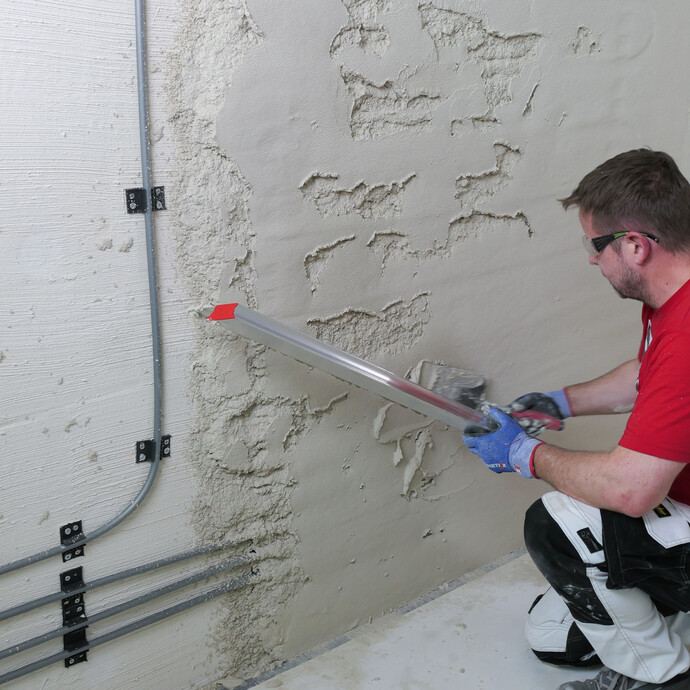 Mann bessert ungleichmässig verputzte Stellen an der Wand mit Putzmaterial und Abziehlatte aus.