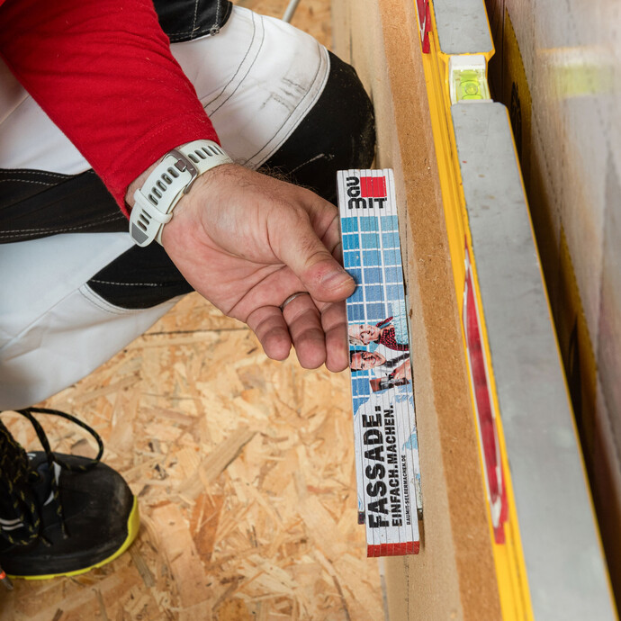 Heimwerker prüft mit Baumit Meterstab als Richtlatte, ob Schraubdübel oberflächenbündig mit der Holzfaserdämmplatte abschließen. Kontrolle bei der Innendämmung.