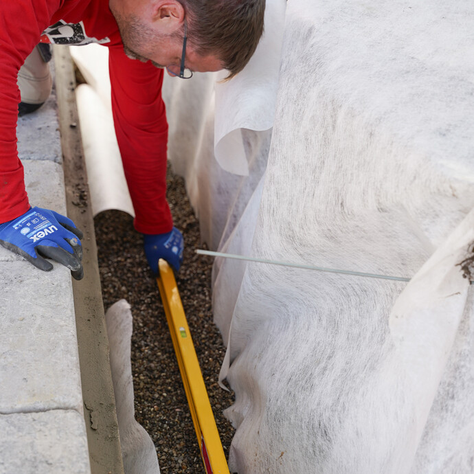 Mit einer langen Wasserwaage wird das Kiesbett für die Drainageleitung ausgerichtet.