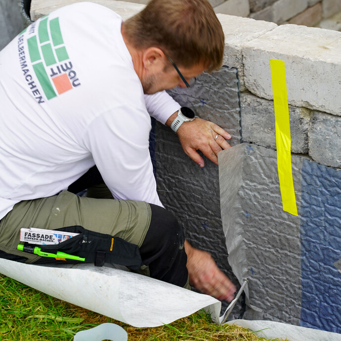 Der Handwerker verklebt die beiden anstoßenden Noppenbahnen miteinander an der Gartenmauer. Rechts ist die Noppenbahn mit einem gelben Klebestreifen fixiert.