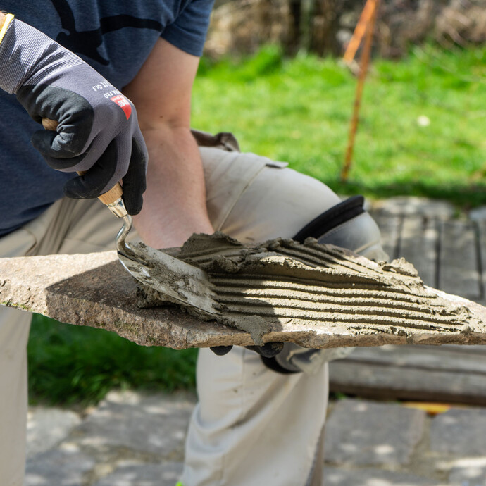 Person trägt mit einer Zahnkelle den Steinkleber auf der Rückseite der zu verklebenden Steinplatte auf.