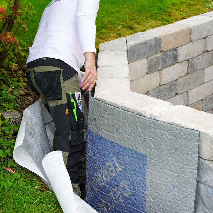 Handwerker setzt die Noppenbahn an der Gartenmauer an. Auf der linken Seite ist ein Hang mit grüner Wiese.