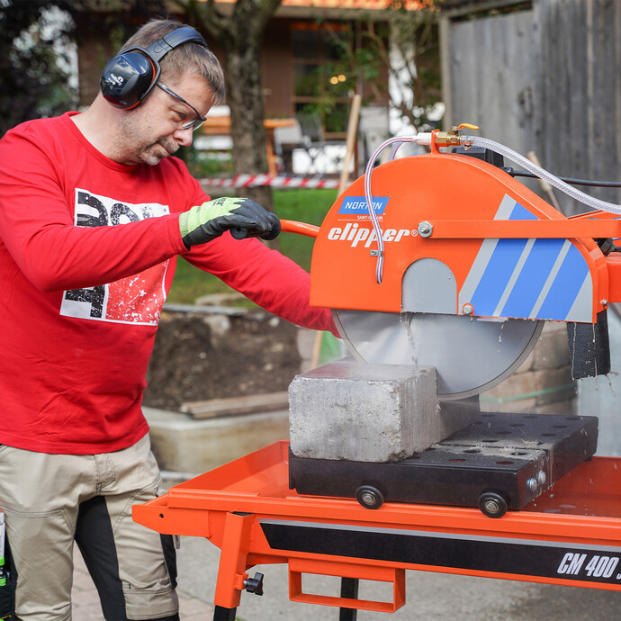 Heimwerker sägt Natursteine und schneidet Betonsteine im Garten mit der Norton Clipper Steinsäge, um eine Gartenmauer zu bauen. Dabei trägt er als Arbeitsschutz einen Gehörschutz und Schnitthandschuhe und Sicherheitsbrille.