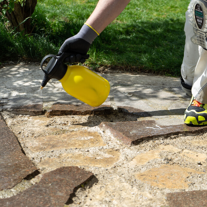 Person sprüht Wasser mit einem gelben Pumpsprüher auf die vorberietet Betonplatte der Terrasse, damit danach die Steinplatten verklebt werden können.