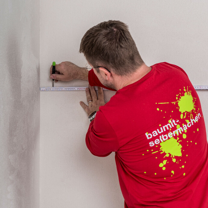 Heimwerker markiert mit Zollstock und Stift die richtige Position für das Baumit Fugendichtband im Stossbereich an der Innenwand – Markierungspunkte setzen für eine bündige Verklebung der Holzfaserdämmplatten, Baumit Selbermachen Anleitung.