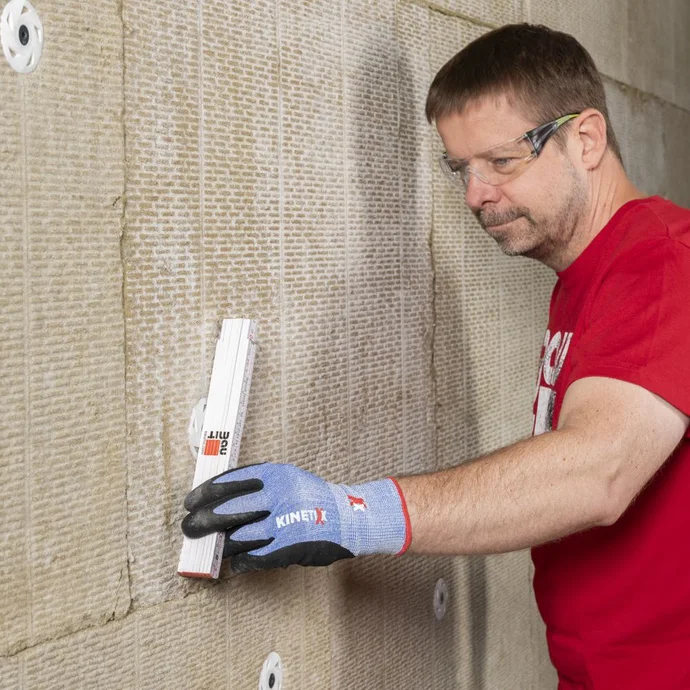 Un homme pose des panneaux isolants sur un mur ; il porte des lunettes de protection et des gants, et montre l'étape de l'isolation de la façade.