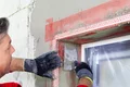Heimwerker glättet Außenputz an einer Fensterlaibung mit Spachtel und Schutznetz für optimale Fassadensanierung.