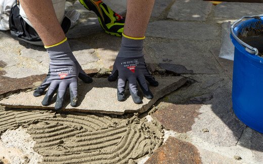 Handwerker drückt eine Natursteinplatte mit Handschuhen in frischen Kleber auf einem gepflasterten Boden fest.