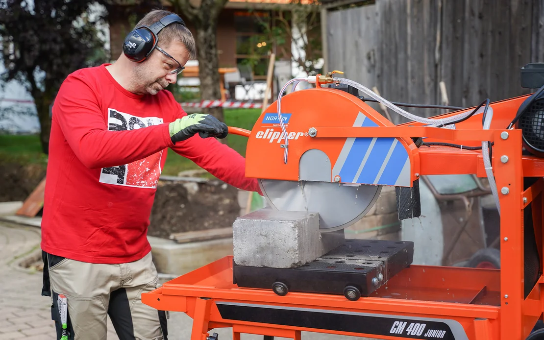 Heimwerker s&auml;gt Natursteine und schneidet Betonsteine im Garten mit der Norton Clipper Steins&auml;ge, um eine Gartenmauer zu bauen. Er tr&auml;gt Geh&ouml;rschutz, Schutzbrille und Arbeitshandschuhe.