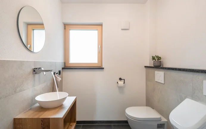 Modernes Gäste-WC mit weissem Waschbecken mit fliessendem Wasser auf Sideboard aus Holz inszeniert mit edlen grauen Wandplatten und wohngesundem Edelputz InStyle Edelweiss in feiner, gefilzter Struktur.