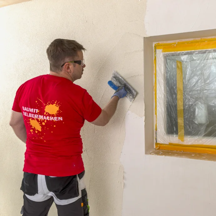 Mann verputzt Innenwand mit Glättkelle, Fenster mit Folie abgedeckt, Heimwerkerprojekt mit Baumit-Produkten.