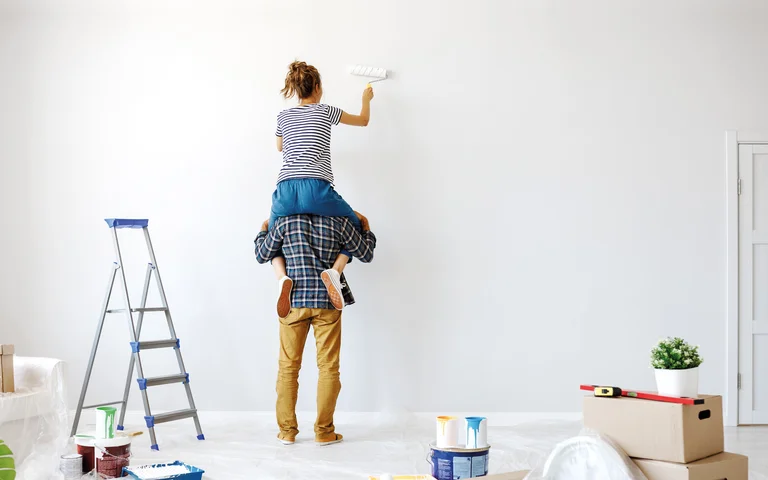 Couple en train de peindre un mur blanc à l’aide de rouleaux à peinture ; projet de rénovation à domicile ; seau et échelle au premier plan.