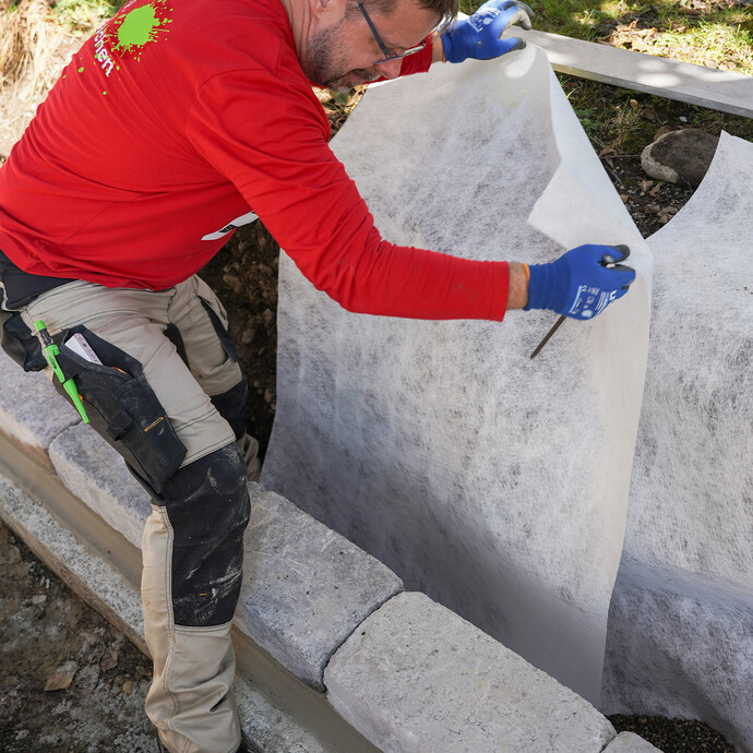 Heimwerker legt ein Drainagevlies in die Baugrube im Garten, damit die Kiesschicht und die Drainageleitung nicht verschlämmt wird.
