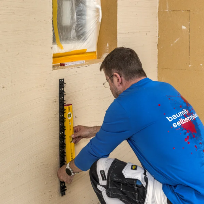 Mann misst Wand mit Wasserwaage, um Fensterrahmen präzise einzubauen, vorbereitet mit Baumaterialien von Baumit.
