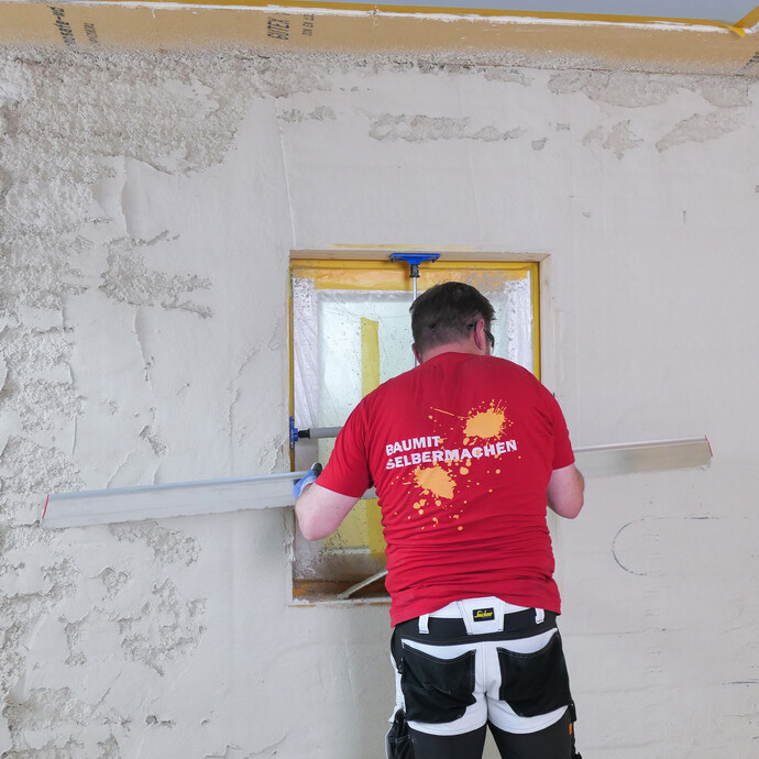Heimwerker glättet Putz mit einer Kartätsche waagrecht entlang einer Putzlehre an einem Fenster.