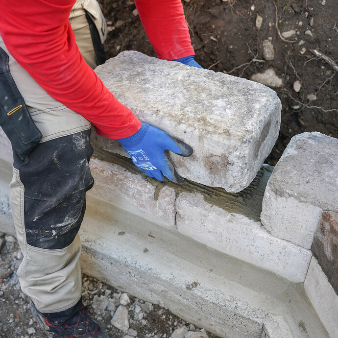 Person platziert einen schweren Mauerstein in den zuvor aufgetragenen und aufgezahnten Steinkleber.