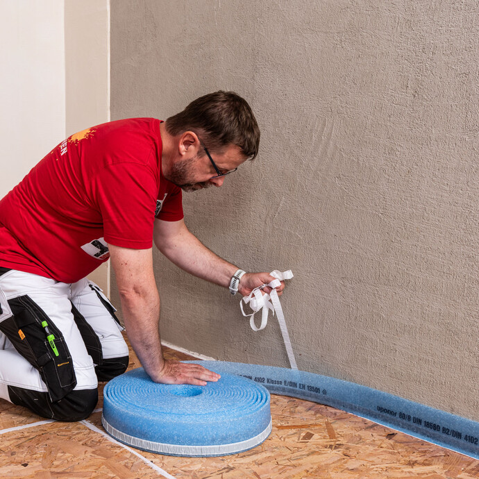 Heimwerker befestigt den Randdämmstreifen an der Wand. Er zieht die Folie des selbstklebenden Dämmstreifens parallel ab.