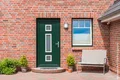 Rote Klinkerfassade mit grüner Haustür und Fenster, begleitet von Pflanzen und einer Bank, ideal für DIY-Renovierungsprojekt.
