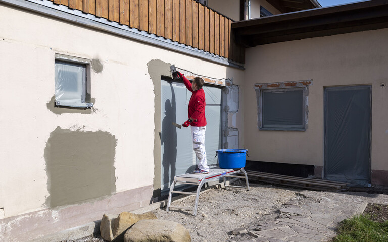 Mann steht auf einem Gerüst und verputzt das Kantenprofil und die Hohlstellen oberhalb der Terrassentüre. Daneben steht ein blauer Mörtelkübel mit den Fassadenspachtel.