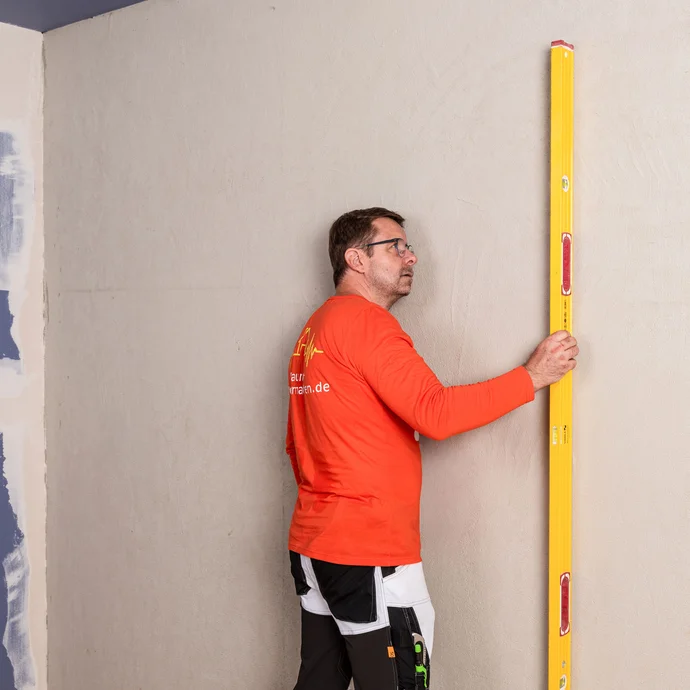 Heimwerker prüft mit einer langen Wasserwaage, ob die Wand eben ist. Eine ebene Wand ist die Voraussetzung für die Montage der Innendämmung.