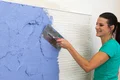Une femme applique de l'enduit bleu au mur à l'aide d'une truelle, projet de bricolage avec des produits Baumit, instructions étape par étape pour la décoration murale.