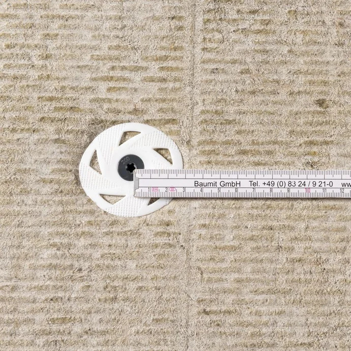 Un mètre ruban mesure l’épaisseur d’un isolant ; on aperçoit une rondelle de fixation sur le matériau de construction.