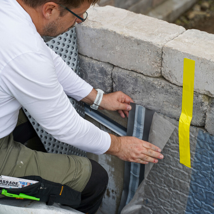 Der Handwerker knickt das Vlies der Noppenbahn zur Seite und entfernt den Schutzstreifen, um die beiden Bahnen im Stoßbereich zu verkleben. Ein gelbes Klebeband fixiert provisorisch die Noppenbahn an der Gartenmauer.