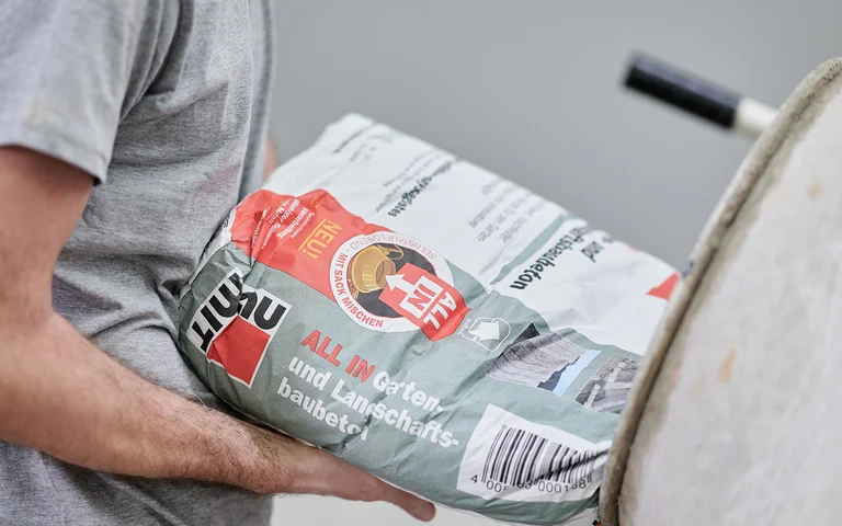 Un bricoleur tient un sac de matériaux Baumit et prépare le matériel pour un projet de jardinage ou d’aménagement paysager.