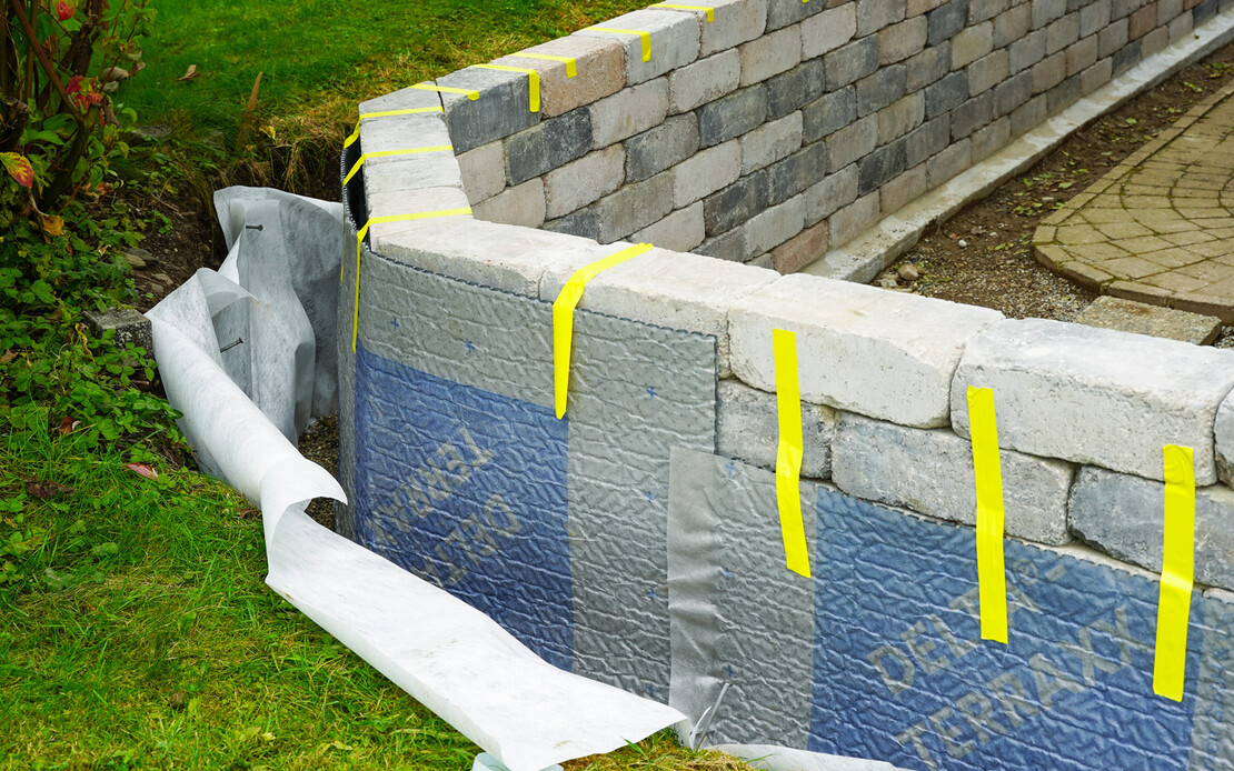 Die Noppenbahn mit Vlies ist provisorisch im Garten an der Gartenmauer mit gelben Klebebandstreifen fixiert. Links davon ist das Geovlies mit Filterkies im erdber&uuml;hrten Bereich zu erkennen. Direkt daneben ist Wiese zu sichtbar.