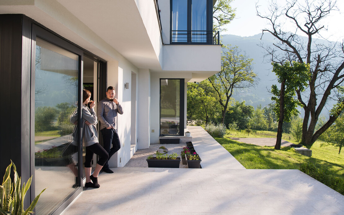 Ein junges Paar trinkt im Stehen auf ihrer Terrasse Kaffee und genie&szlig;t die Sonne.