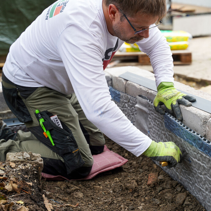 Nachdem die Baugrube wieder aufgefüllt wurde, kürzt der Handwerker mit einem Cuttermesser die Noppenbahn entlang der unteren Kante des Abschlussprofils.