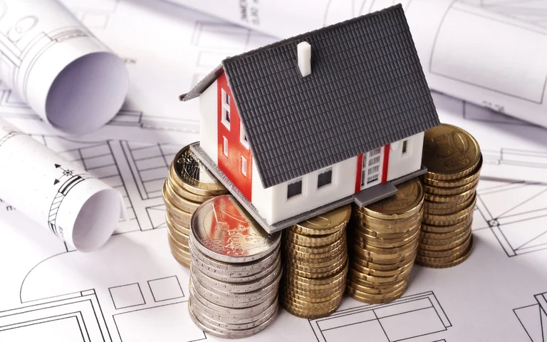 Miniaturhaus auf Münzstapeln, symbolisiert Baufinanzierung und Budgetplanung für Heimwerkerprojekte, mit Bauplänen im Hintergrund.