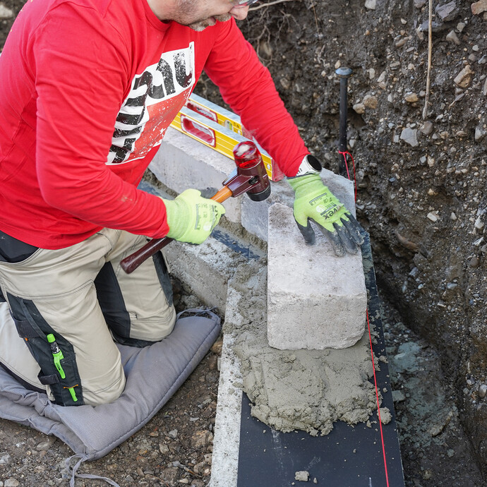 Eine Person richtet einen gesetzten Mauerstein während des Baus einer Gartenmauer mit einer Wasserwaage und Hammer aus.