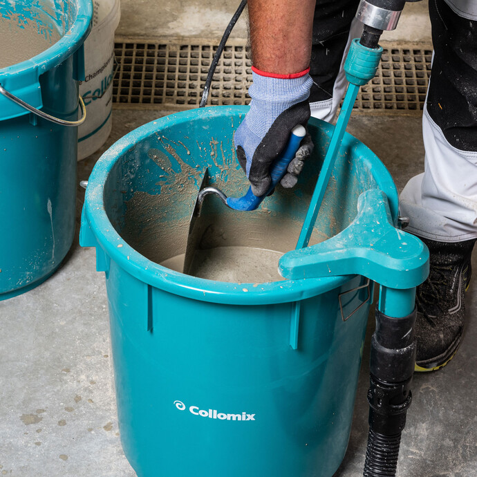Photo en gros plan : Seau bleu contenant du mortier d'égalisation mélangé. Personne raclant les restes de mortier sur le bord du seau à l'aide d'une truelle.