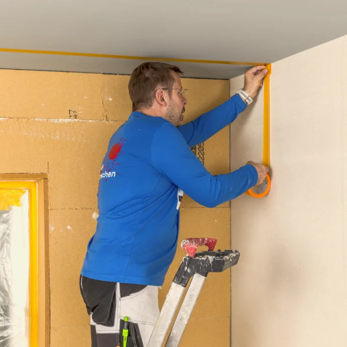 Heimwerker klebt gelbes Malerkrepp an die Wand, bereitet Renovierungsarbeiten vor. Leitern und Abdeckmaterial im Hintergrund.