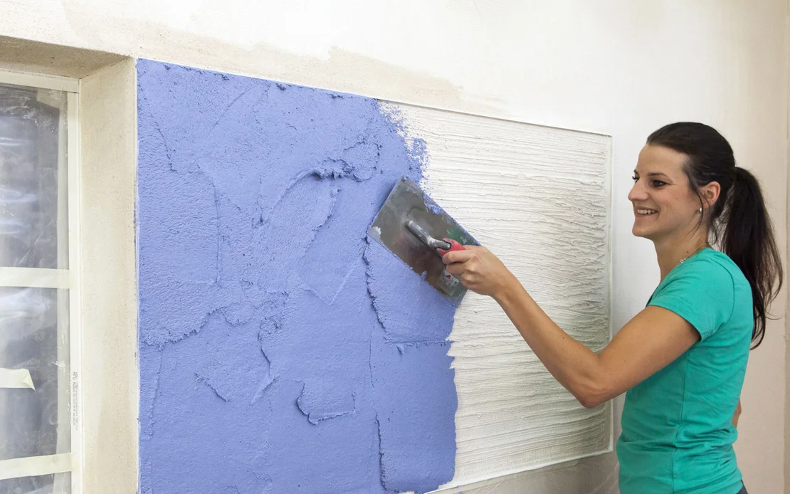 Frau tr&auml;gt blauen Kratzputz mit einer Gl&auml;ttkelle auf die vorbereitete Fassade auf.