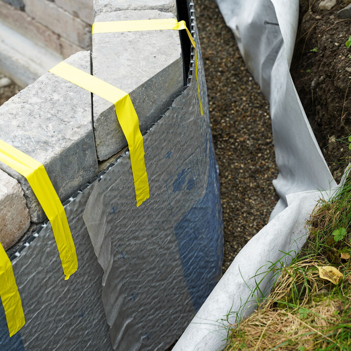 Die Noppenbahnen an der Gartenmauer sind provisorisch mit Klebeband fixiert, damit sie in ihrer Position nicht verrutschen. Rechts unten ist das Geovlies mit Filterkies zu erkennen.