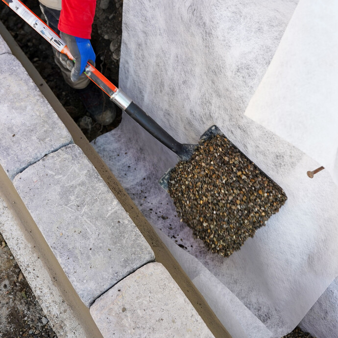 Handwerker mit blauen Arbeitshandschuhen schüttet mit einer Schaufel Filterkies auf das Geovlies.