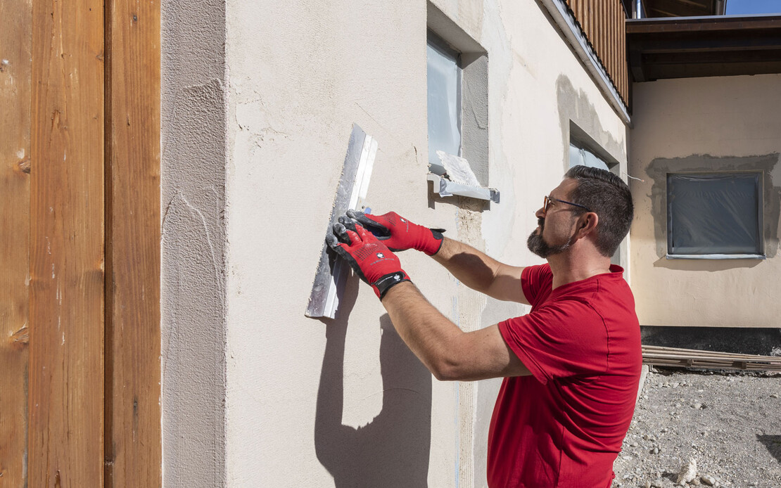 Mann glättet den aufgetragenen Putz mit einer Flächenspachtel an der Fassade.