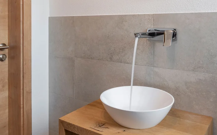 Modernes, weisses Waschbecken mit fliessendem Wasser auf Sideboard aus Holz inszeniert mit edlen grauen Wandplatten mit wohngesundem Edelputz in gefilzter Struktur.
