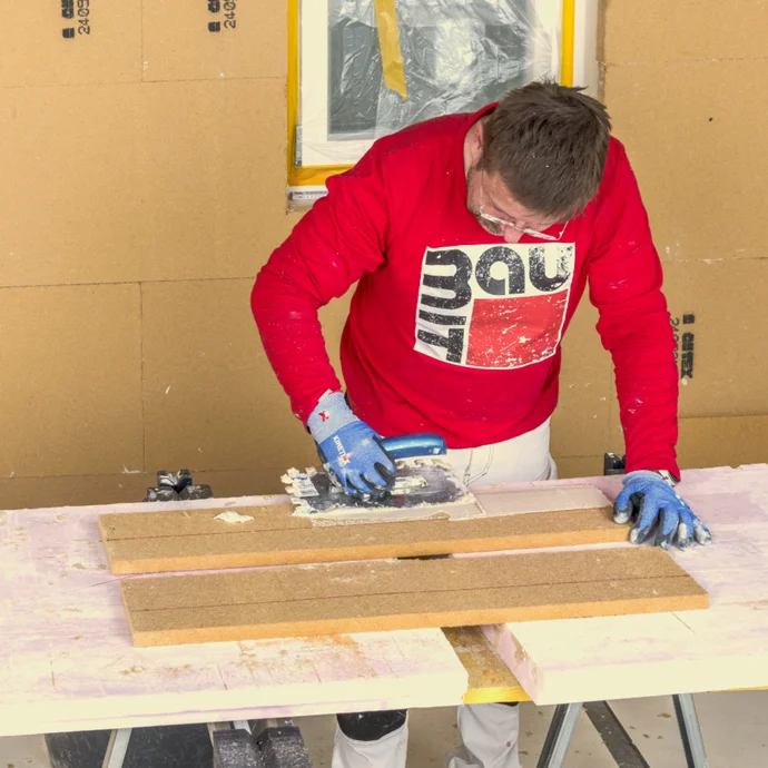 Un bricoleur découpe un panneau isolant sur un établi, vêtu d’un sweat-shirt Baumit.