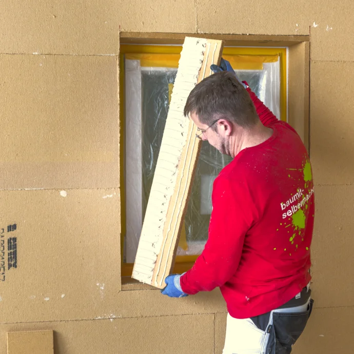 Un homme isole des châssis de fenêtre avec des panneaux isolants, vêtu d’une veste Baumit rouge, dans le cadre d’une rénovation d’intérieur.