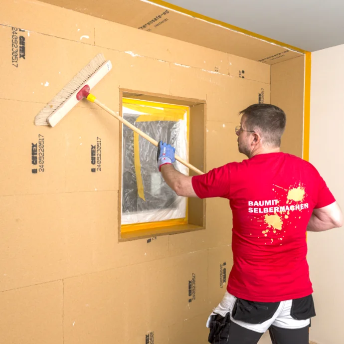 Heimwerker glättet Trockenbauwand mit Besen in renoviertem Raum, um Oberflächen für weitere Arbeiten vorzubereiten.