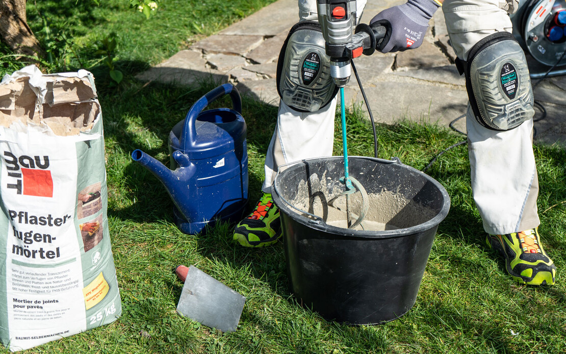 Person r&uuml;hrt mit einem elektrischen R&uuml;hrger&auml;t den Pflasterfugenm&ouml;rtel im Garten an.