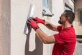 Mann verputzt Außenwand mit Glättkelle, trägt rote Handschuhe, Tageslicht.