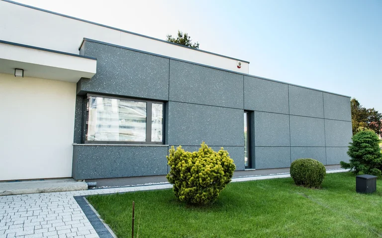 Façade moderne d’une maison individuelle avec un enduit extérieur gris de la marque Baumit, entourée d’une pelouse bien entretenue et d’arbustes.