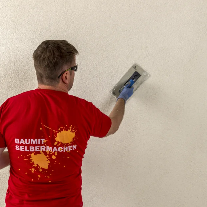 Heimwerker beim Auftragen von Putz auf eine Wand mit einer Glättkelle, im roten Baumit-Shirt.