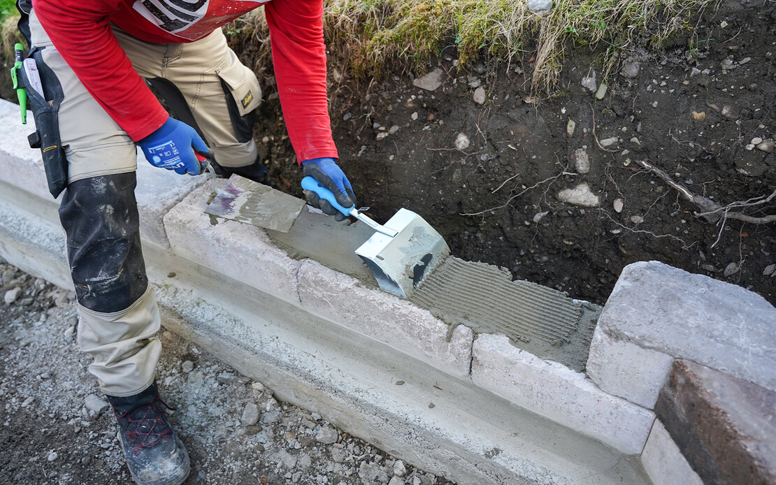 Natursteine und Betonsteine kleben mit Baumit Steinkleber Trass - Mit einer Klebekelle wird der aufgetragene Steinkleber aufgeraut, um eine bessere Haftung zu gew&auml;hrleisten. So kann der n&auml;chste Mauerstein der Gartenmauer gesetzt werden.