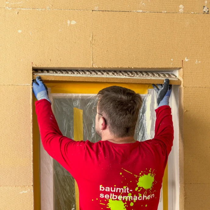 Un bricoleur enduit soigneusement le bord d’un encadrement de porte avec de l’enduit Baumit, en le protégeant d’un film de protection pour un résultat précis.